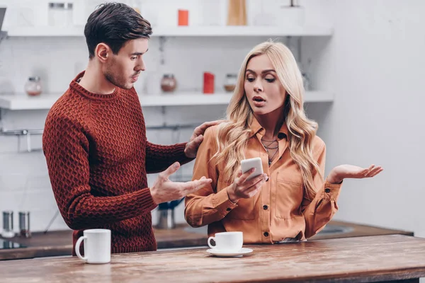 Jealous young man looking at emotional girlfriend using smartphone in kitchen, distrust concept — Stock Photo