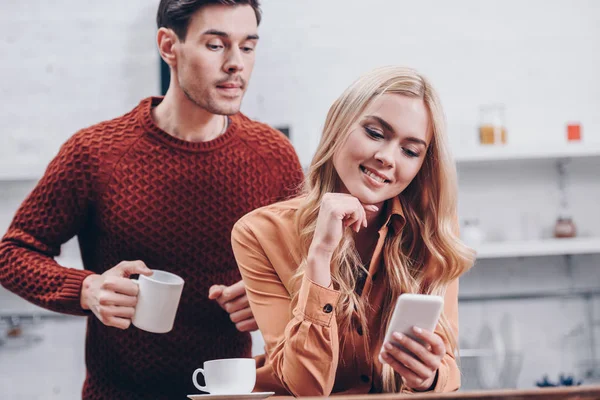 Jealous young man holding cup and looking at smiling girlfriend using smartphone in kitchen, mistrust concept — Stock Photo