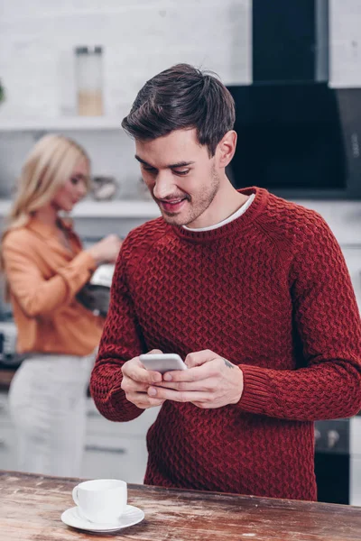 Sorridente giovane uomo sms tramite smartphone mentre la ragazza tiene il bollitore dietro in cucina — Foto stock