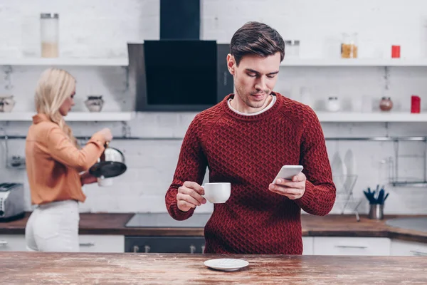 Joven hombre sosteniendo taza y el uso de teléfono inteligente en la cocina, concepto de problema de relación - foto de stock