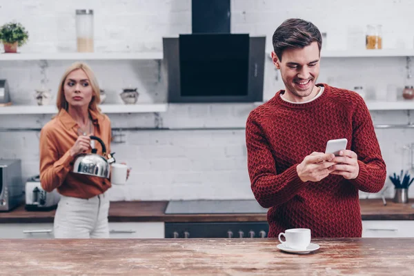 Jealous young woman with kettle looking at smiling husband using smartphone in kitchen — Stock Photo