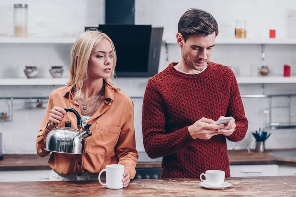 Jealous young woman with kettle looking at husband with smartphone in kitchen — Stock Photo