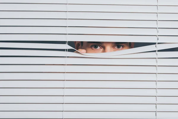 Joven sospechoso espiando y mirando a la cámara a través de persianas, concepto de desconfianza - foto de stock