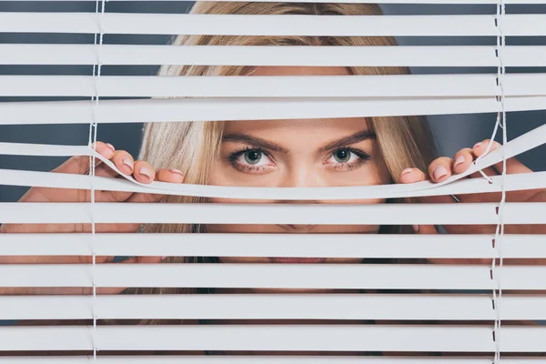 Junge Frau blickt in Kamera und blickt durch Jalousien — Stockfoto