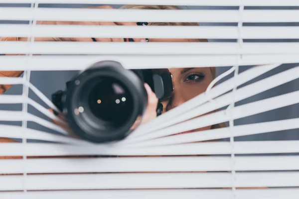 Nahaufnahme von Kamera und junger Frau, die Fotos macht und durch Jalousien spioniert — Stockfoto