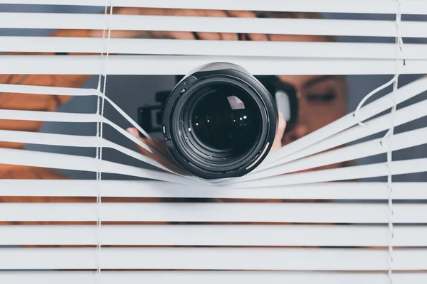 Close-up view of camera and young woman spying through blinds — Stock Photo