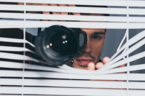 Jovem homem segurando câmera e olhando para a câmera através de persianas, conceito de desconfiança — Fotografia de Stock