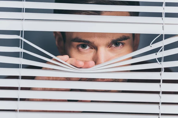 Verdächtiger junger Mann blickt durch Jalousien in Kamera, misstraut Konzept — Stockfoto