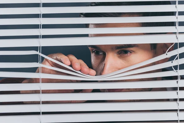 Jeune homme méfiant regardant loin et regardant à travers les stores — Photo de stock