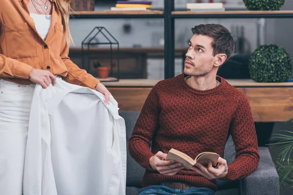 Corte tiro de mulher ciumenta mostrando camisa branca com traços de batom para marido segurando livro em casa — Fotografia de Stock