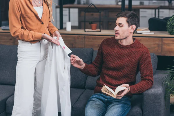 Cropped shot of jealous woman showing male shirt with lips traces to husband holding book at home — Stock Photo