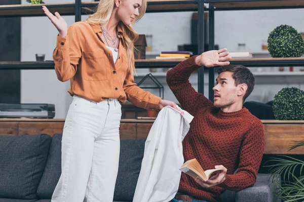 Corte tiro de mulher com ciúmes raiva mostrando camisa masculina com lábios traços de homem assustado segurando livro em casa — Fotografia de Stock