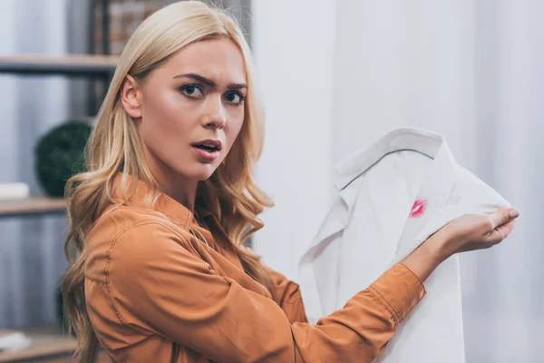 Upset young woman holding white male shirt with lipstick traces and looking at camera, betrayal concept — Stock Photo