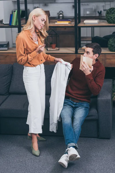 Celosa mujer mostrando camisa masculina con labios rastros de hombre asustado sosteniendo libro en casa - foto de stock