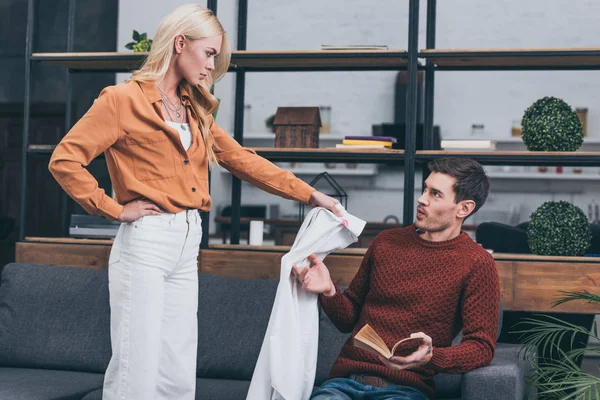 Angry young woman showing male shirt with lipstick traces to confused man reading book — Stock Photo