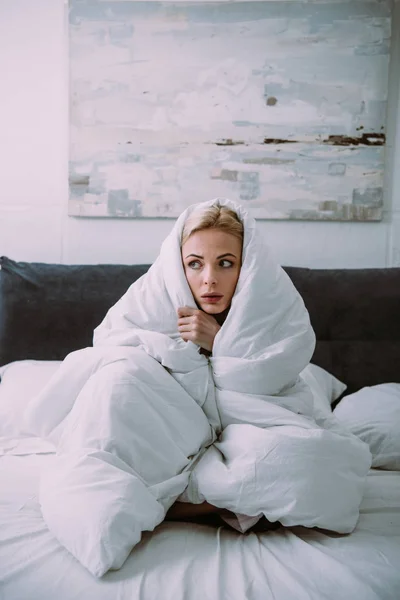 Испуганная женщина, покрытая одеялом, смотрит в кровать — стоковое фото