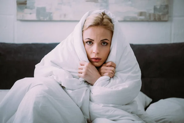Frightened woman covered in blanket looking at camera in bed — Stock Photo