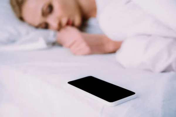 Selective focus of smartphone with blank screen and woman lying in bed on background — Stock Photo