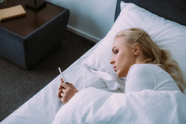 Femme seule couchée au lit et utilisant un smartphone à la maison — Photo de stock