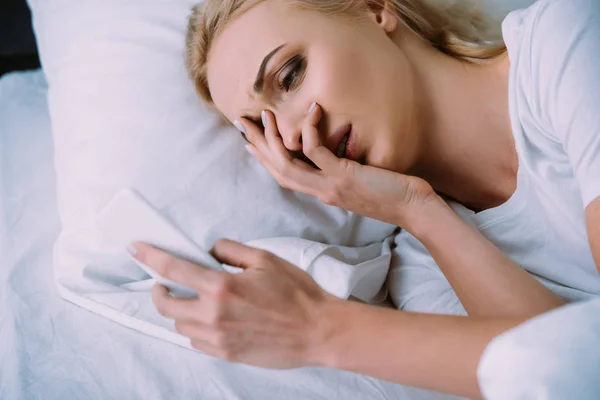 Donna sconvolta sdraiata a letto, che copre il viso con la mano e utilizzando lo smartphone — Foto stock