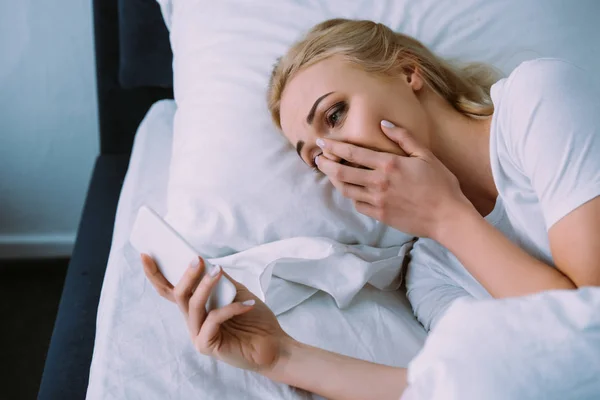 Verärgerte Frau liegt im Bett, deckt Gesicht mit der Hand ab und benutzt Smartphone — Stockfoto