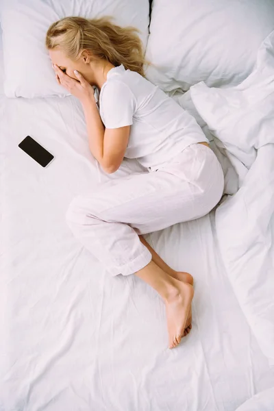Vista dall'alto della donna che copre il viso e piange mentre giace vicino allo smartphone con schermo vuoto a letto a casa — Foto stock
