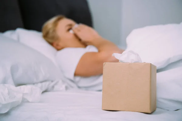 Selektive Fokussierung der Gewebebox mit einer Frau, die Tränen im Bett wischt — Stockfoto