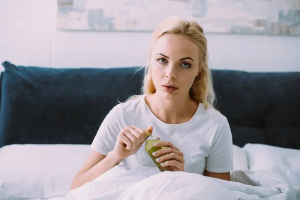 Засмучена жінка дивиться на камеру і тримає пляшку з ліками в ліжку — стокове фото
