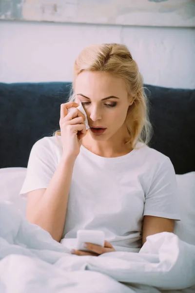 Crying woman wiping tears and using smartphone in bed at home — Stock Photo