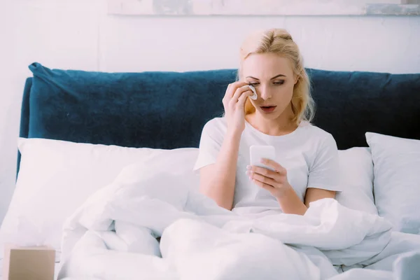 Crying woman wiping tears and using smartphone in bed at home — Stock Photo