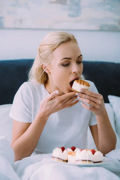 Schöne traurige Frau im weißen Pyjama, die Kuchen allein im Bett isst — Stockfoto