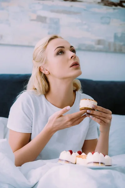 Upset woman in white pajamas holding cake in bed alone and looking away — Stock Photo