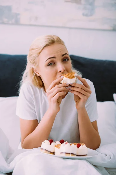 Mujer molesta en pijama mirando a la cámara y comiendo pastel en la cama solo - foto de stock