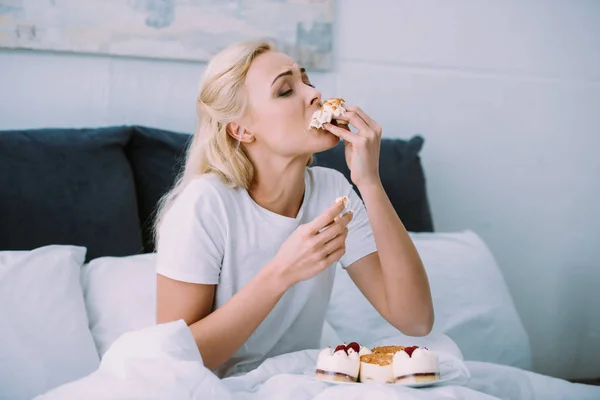 Stressed woman in pajamas eating sweet cake in bed alone — Stock Photo
