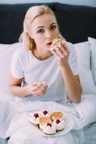 Bouleversé femme en pyjama regarder caméra et manger gâteau dans le lit seul — Photo de stock