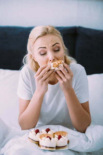 Stressed woman in pajamas eating cake in bed alone — Stock Photo