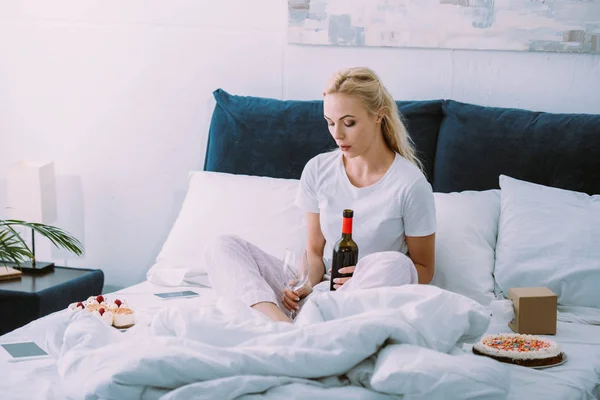 Donna triste con bicchiere e bottiglia di vino che festeggia il compleanno a letto da sola — Foto stock