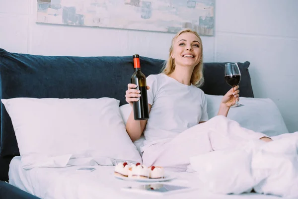 Donna sorridente che tiene bottiglia e bicchiere di vino rosso mentre festeggia il compleanno a letto da solo — Foto stock