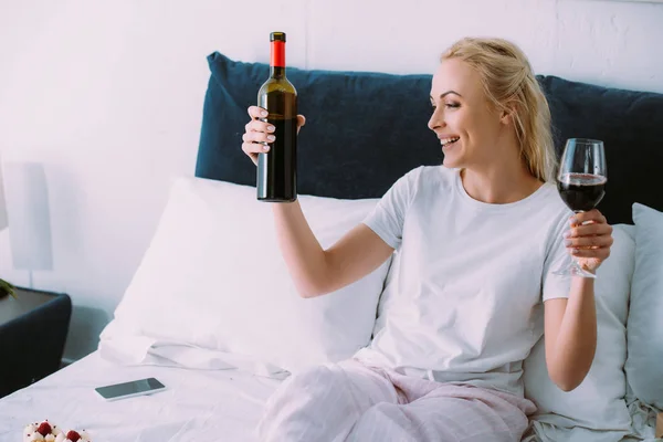 Hermosa mujer sonriente en pijama sosteniendo botella de vino y vidrio en la cama en casa - foto de stock