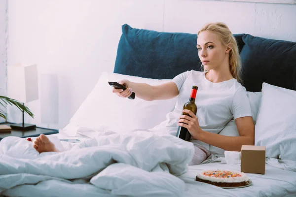Sad woman with wine bottle watching tv while celebrating birthday in bed alone — Stock Photo