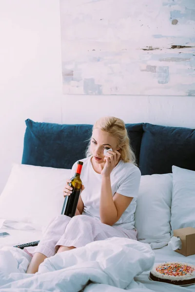 Chateado mulher com garrafa de vinho enxugando lágrimas enquanto comemora aniversário na cama sozinho — Fotografia de Stock