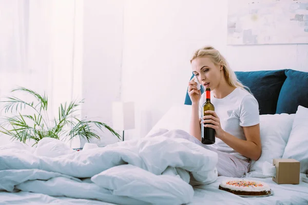 Donna triste con bottiglia di vino asciugare le lacrime mentre festeggia il compleanno a letto da solo — Foto stock