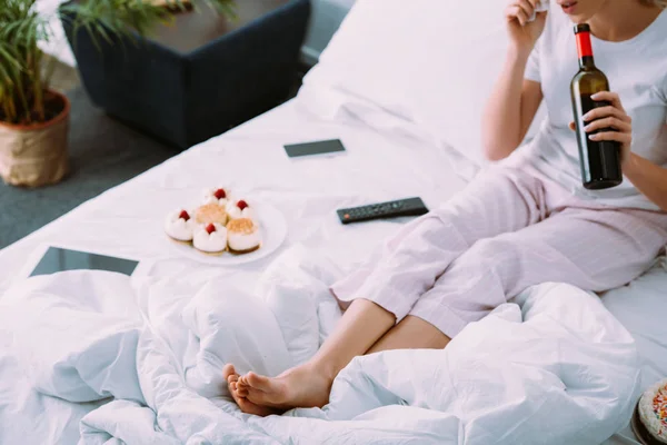 Cropped view of woman with cakes and bottle of wine celebrating birthday in bed alone — Stock Photo