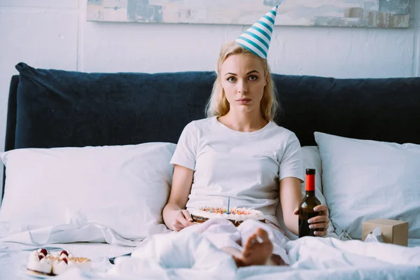 Mulher deprimida com garrafa de vinho e bolo olhando para a câmera enquanto comemora aniversário na cama sozinho — Fotografia de Stock