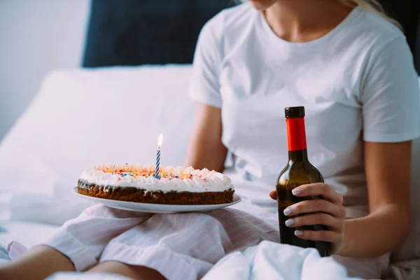 Vista recortada de la mujer con botella de vino y pastel celebrando cumpleaños en la cama solo - foto de stock