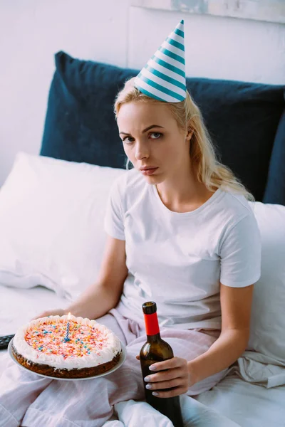 Chateado mulher com garrafa de vinho e bolo comemorando aniversário na cama sozinho — Fotografia de Stock