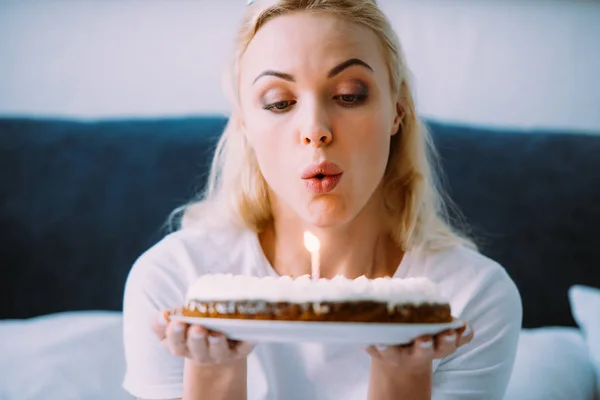 Selective focus of woman blowing out candle on cake while celebrating birthday in bed alone — Stock Photo