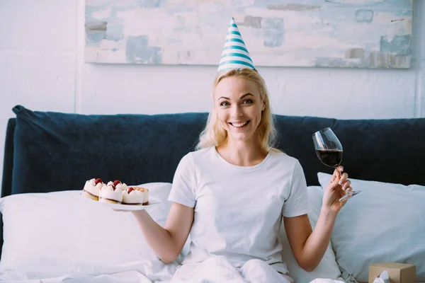 Mulher feliz em chapéu de festa segurando copo de vinho tinto e bolo enquanto celebra aniversário na cama sozinho — Fotografia de Stock