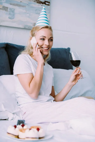 Mulher sorridente em chapéu de festa comemorando aniversário, segurando copo de vinho e falando no smartphone na cama em casa — Fotografia de Stock