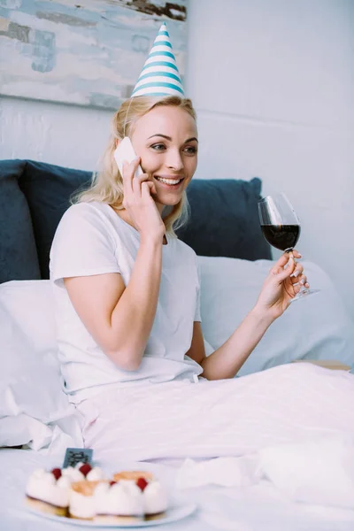 Smiling woman in party hat celebrating birthday, holding glass of wine and talking on smartphone in bed at home — Stock Photo